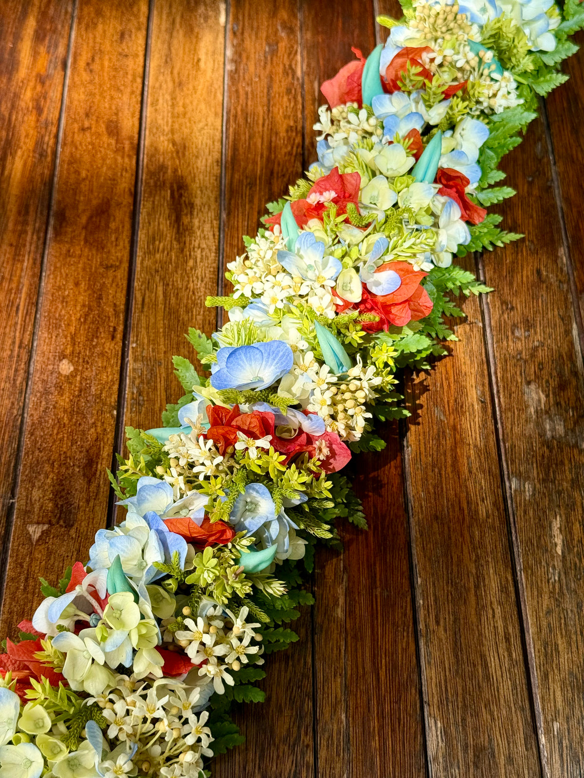 A long floral garland with blue hydrangeas, red flowers, white blossoms, and green foliage laid diagonally across a dark brown wooden plank surface.