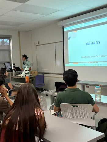A classroom scene with a presenter standing at a desk near a whiteboard and projector screen, while several students sit at desks facing the screen.