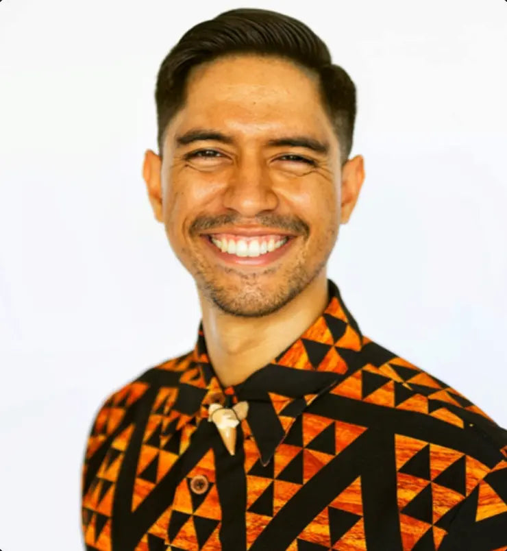 A smiling man with dark hair and a short beard wearing a black and orange geometric patterned button-down shirt, photographed against a plain white background.